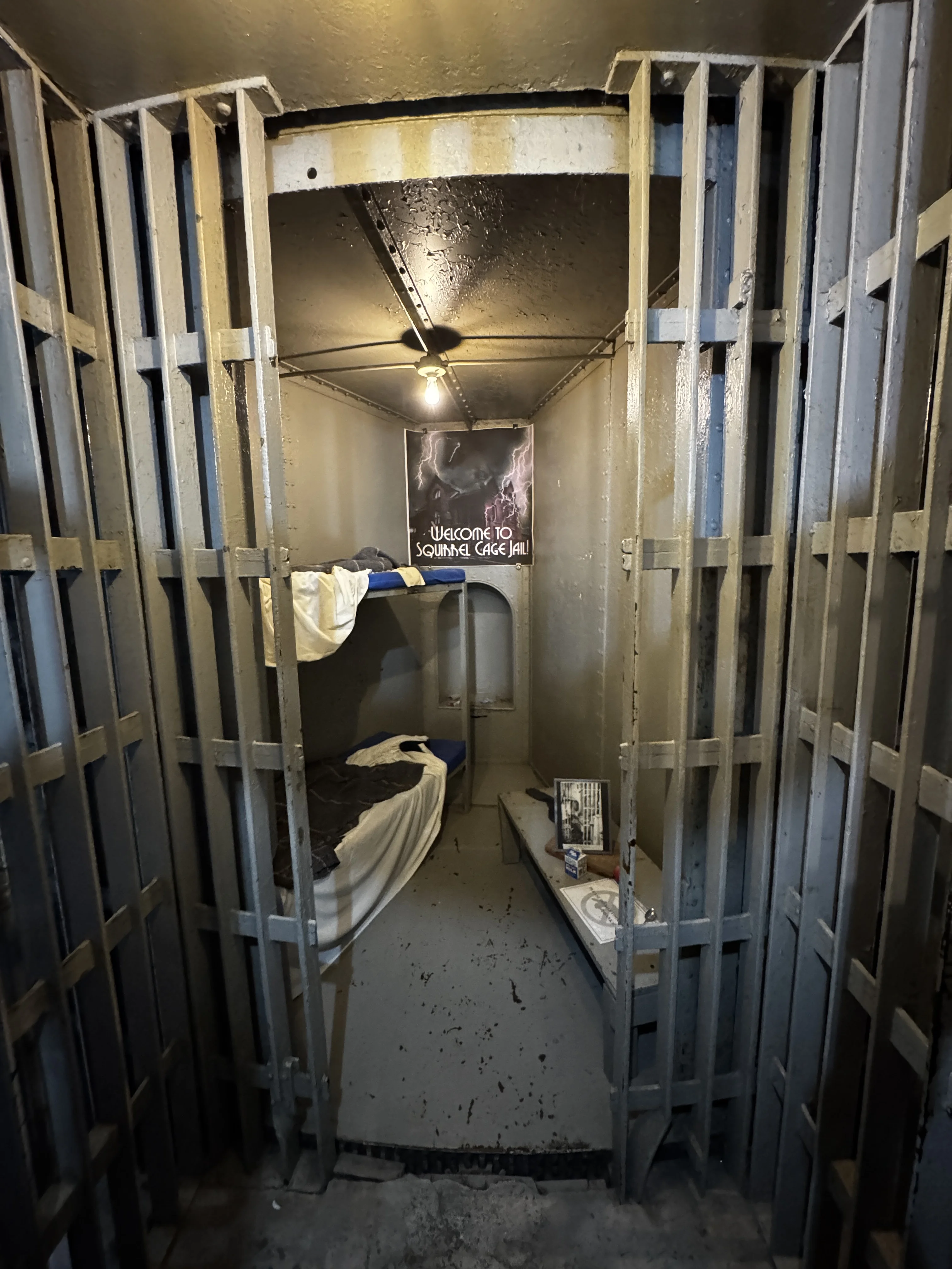 Interior of a pie-slice cell through iron bars, showing narrow tapering steel walls, two bunk beds, and a single ceiling bulb