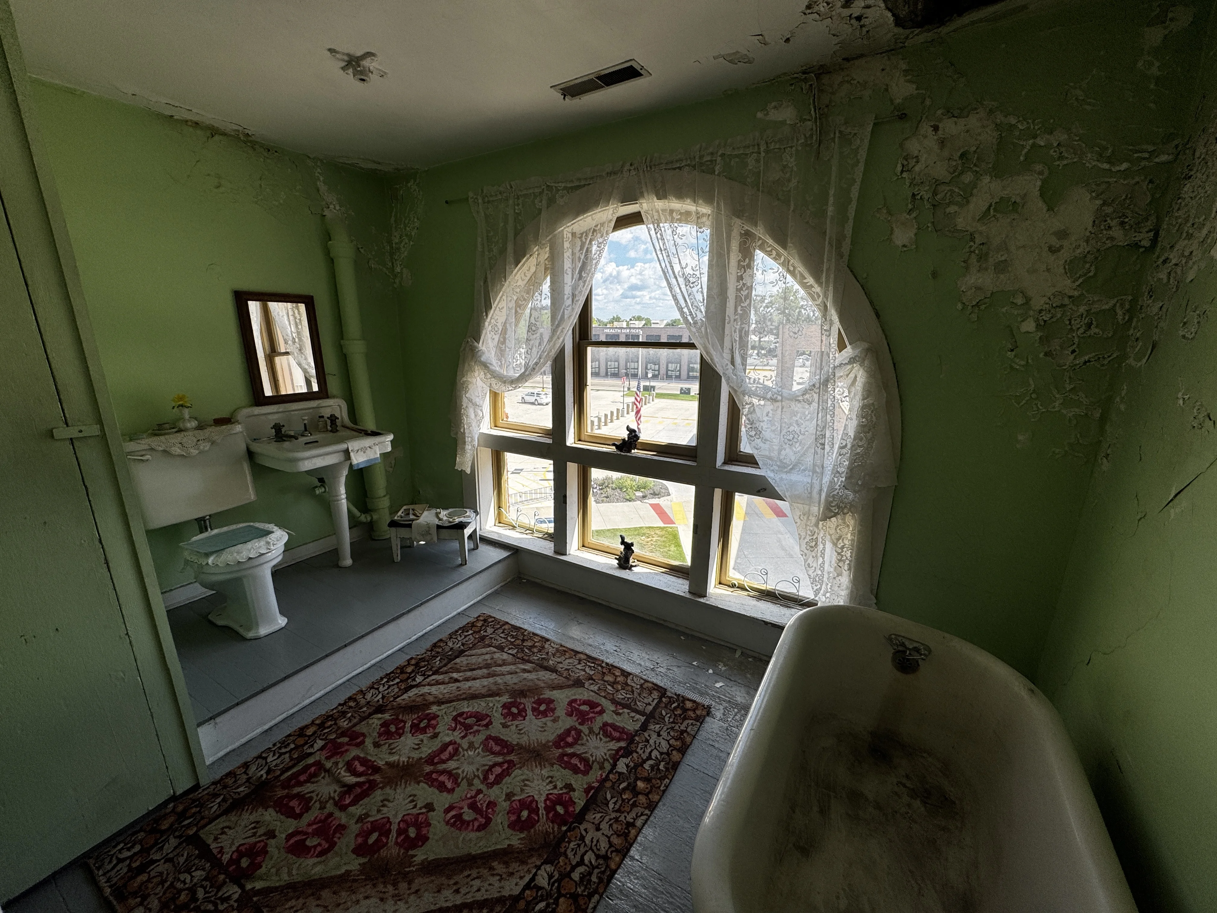 A vintage bathroom with green peeling walls, clawfoot tub, patterned rug, and lace curtains over an arched window overlooking downtown Council Bluffs